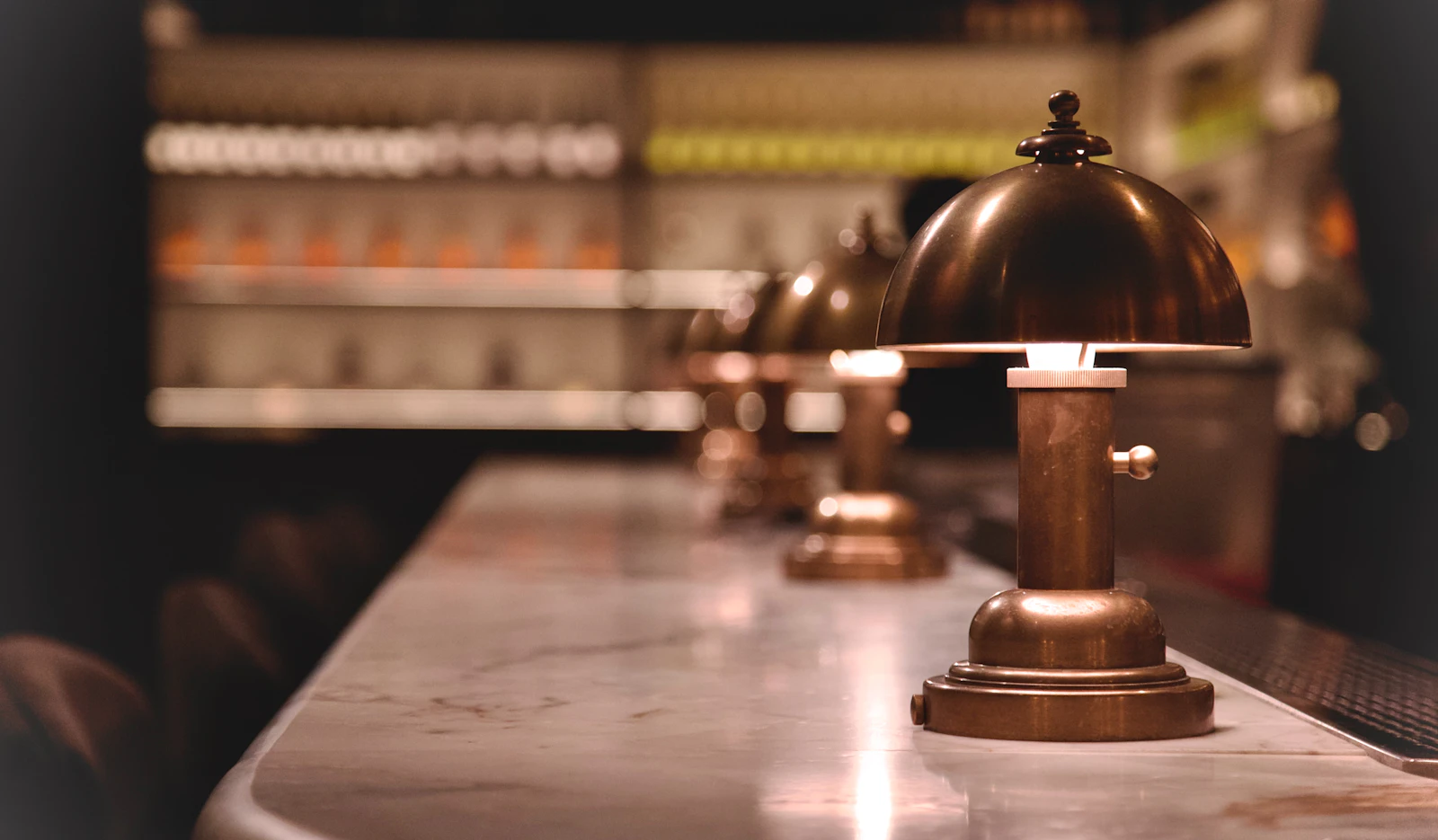 A row of brass table lamps glowing on a marble bar counter, the back bar blurred into warm bokeh behind them.