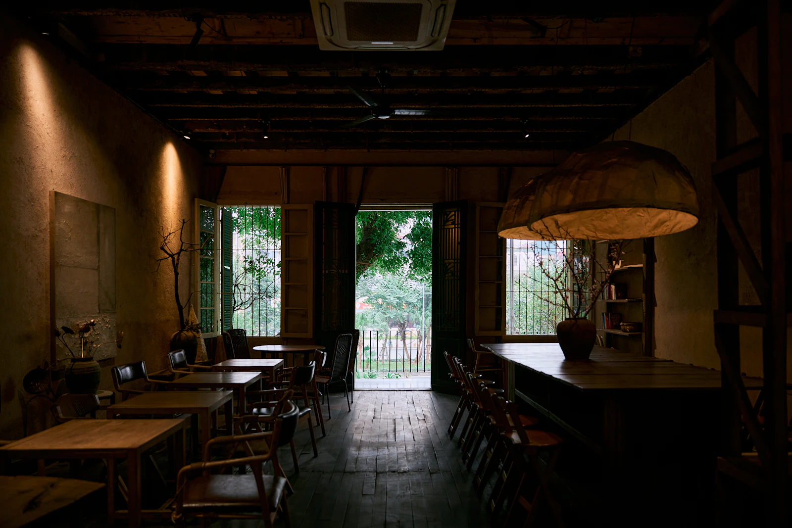 An empty, dimly lit cafe with wooden tables and chairs, a large paper lamp, and open doors onto a sunlit garden.
